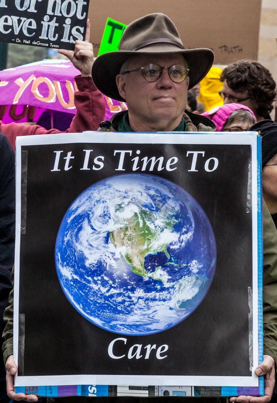 Man in brown felt hat and glasses, at a rally, holding a sign reading "It is Time to Care" with a picture of planet Earth.